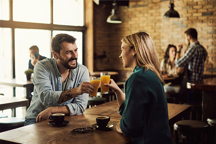 Man and woman clinking glasses of juice in a cozy café, illustrating challenges men face explaining things to women.
