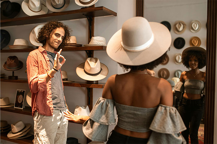 Man and woman in a hat store, man gesturing while woman tries on a hat, illustrating drives my wife crazy moments.