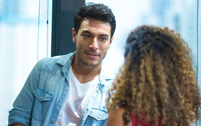 Man in denim shirt talking to woman with curly hair in a cafe, illustrating things men shouldn’t do while pursuing women online. - 6