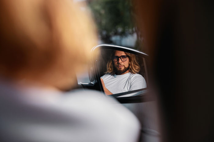 Man with glasses and long hair reflected in car side mirror, illustrating things men shouldn’t do while pursuing women. - 2