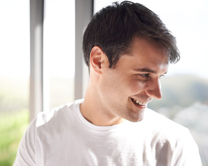 Smiling man in a white t-shirt indoors near window, illustrating things men shouldn’t do while pursuing women advice. - 33