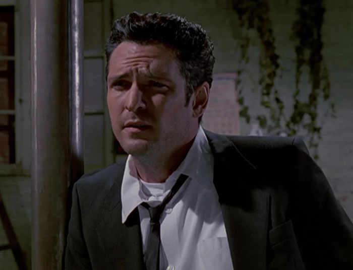 Actor Michael Madsen in a dark suit, looking contemplative with a serious expression in a dimly lit outdoor setting.