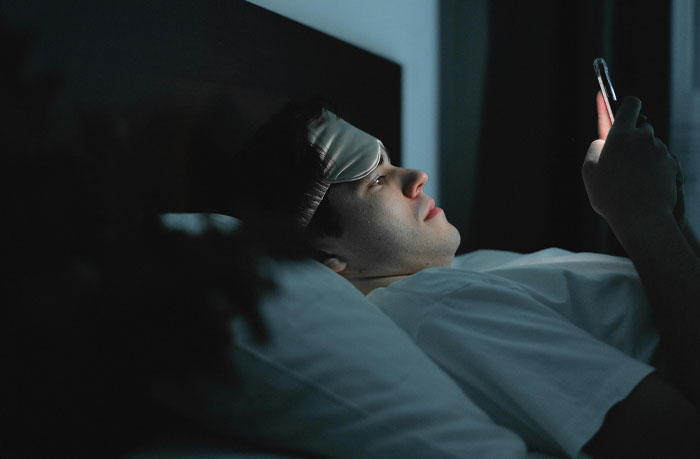 Young man lying in bed at night wearing an eye mask on forehead, looking at phone screen, illustrating micro cheating stories.