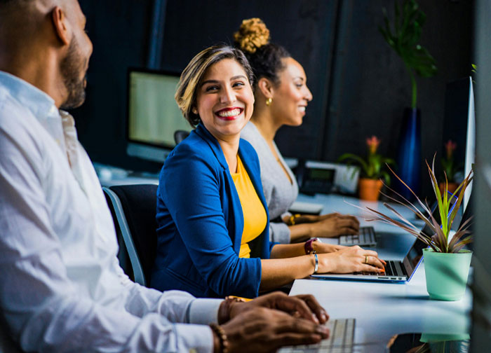 Three coworkers at computers in an office, a woman in blue smiling while another woman and man work, illustrating trust and micro cheating.