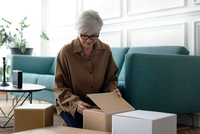 Older woman with glasses opening a package at home, illustrating a nosy MIL who can’t stop opening others’ packages. Older woman with glasses opening a package at home, illustrating a nosy MIL who can’t stop opening others’ packages.