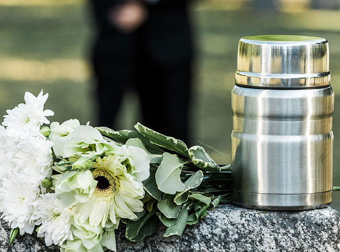 Silver urn on a tombstone next to white flowers with blurred figure in the background, relating to grandson ashes family drama.