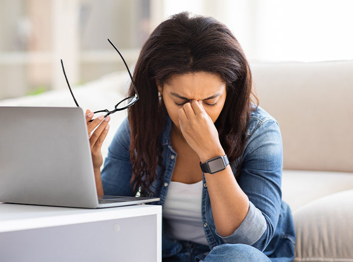 Frustrated woman sitting by laptop, holding glasses and rubbing her eyes, reflecting family drama over grandson&rsquo;s ashes.