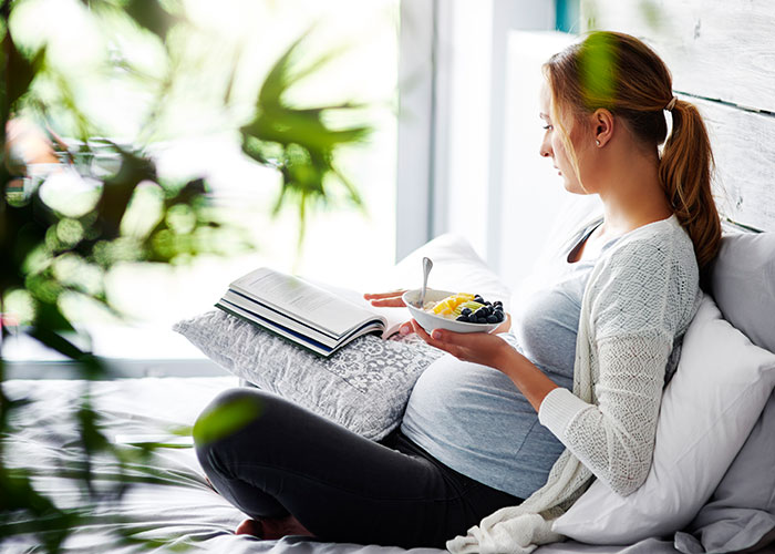 Pregnant woman sitting on bed, holding a bowl of fruit and reading a book, calm before a gender reveal event.