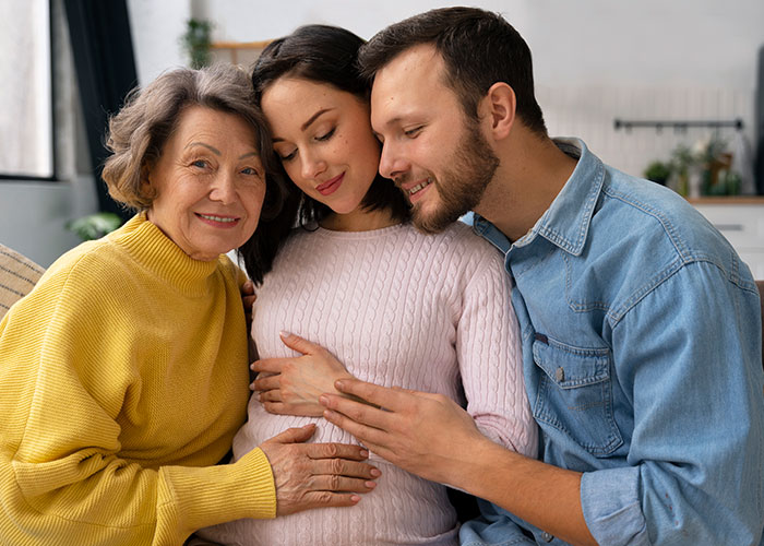 Pregnant woman with partner and mother-in-law touching belly during a gender reveal moment at home.