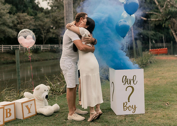 Couple hugging at outdoor gender reveal party with blue smoke indicating boy and balloons in background.