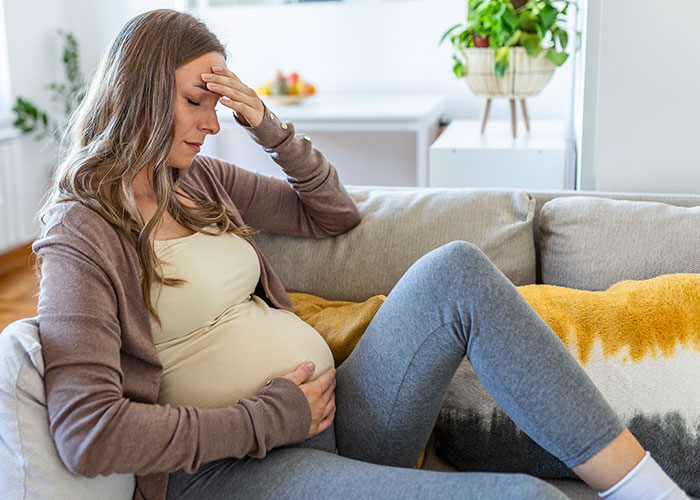 Pregnant woman sitting on couch, looking stressed and holding her belly, reflecting tension at gender reveal event.