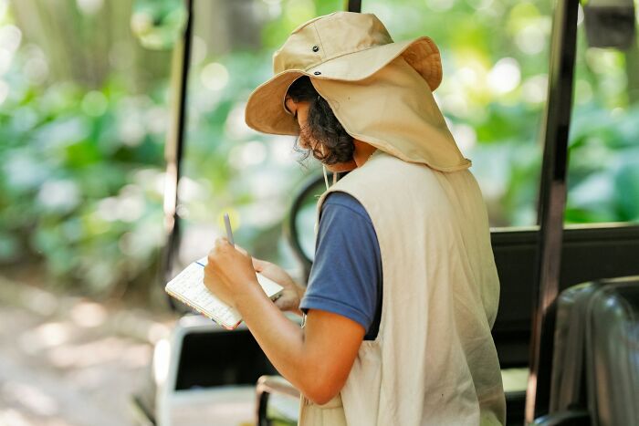Person with a sun hat and vest writing notes outdoors, capturing hard-to-believe stories about themselves in nature.