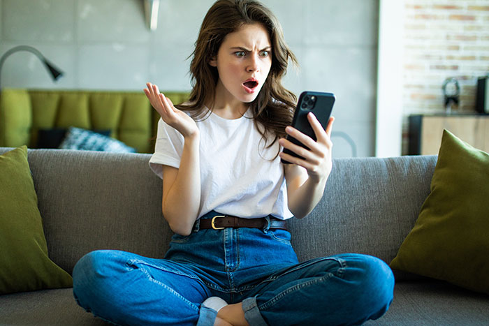 Young woman sitting on a couch looking shocked at her phone, reacting to friends who finally noticed total jerks behavior.
