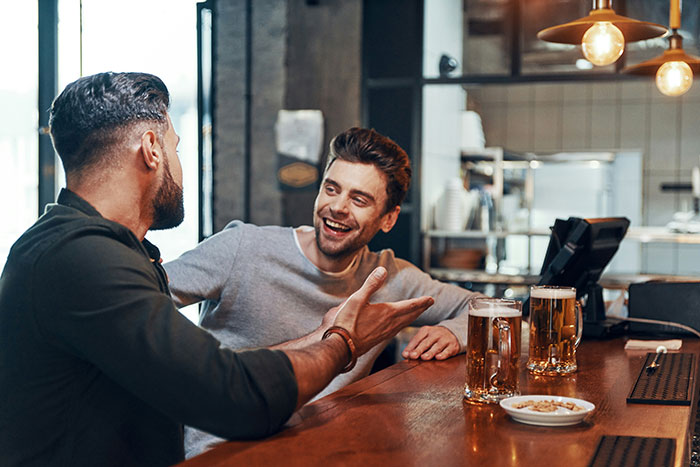 Two friends chatting and laughing at a bar with beers, capturing moments of friendship and realization of true character.