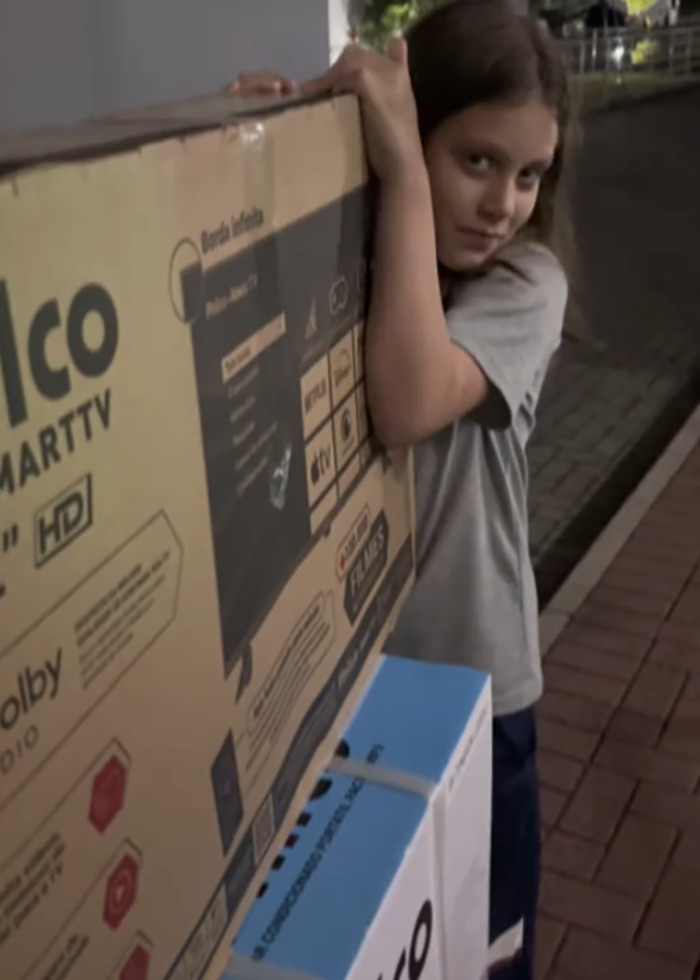 Young girl carries large TV box outdoors, illustrating momfluencer controversy involving autistic daughter and balcony sleeping arrangement.