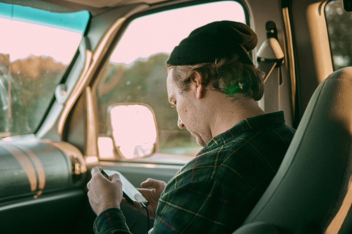 Man wearing a beanie and plaid shirt sitting in a car, looking down at his phone, reflecting on generational trauma discussion. - 1