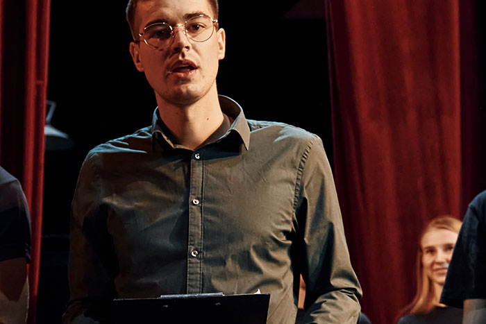 Young man wearing glasses and a dark shirt speaking on stage during a classic school moment event with red curtains.
