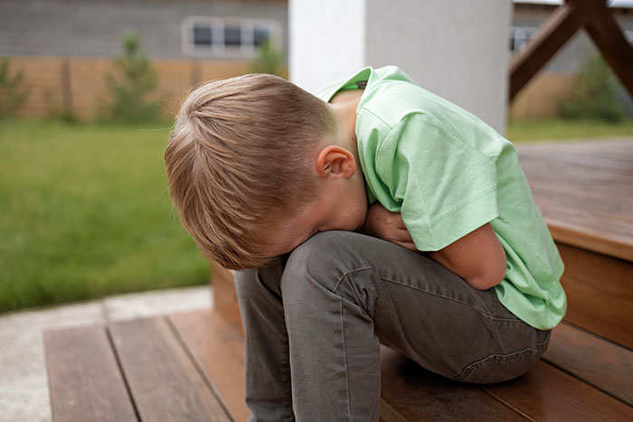 Young boy in green shirt sitting on steps with head down, experiencing a cringeworthy school moment in an outdoor setting.