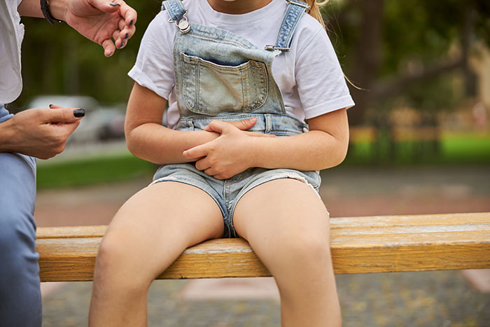 Child sitting on bench holding stomach while an adult offers a pill, capturing cringeworthy school moments and embarrassment.