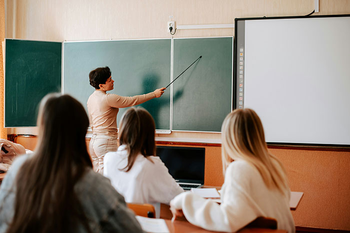 Teacher pointing at chalkboard in a classroom with students watching, capturing classic school moments that cause red-faced reactions.