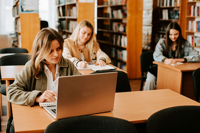 Three female students in a library working on laptops and notes, capturing classic school moments and learning environment.