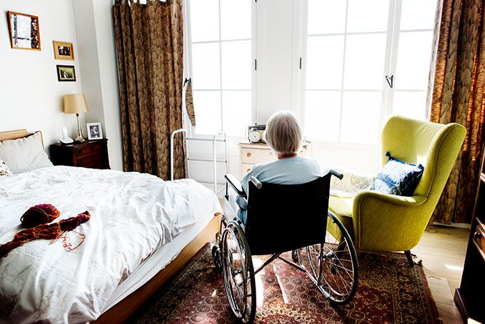 Elderly person in a wheelchair looking out bright bedroom window, evoking eerie and creepy mortician stories.