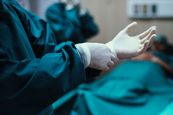 Mortician wearing gloves preparing for embalming with a blurred patient in the background in a clinical setting