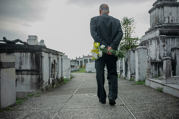 Man in dark suit holding flowers walking through cemetery path, evoking creepy and disturbing mortician stories atmosphere