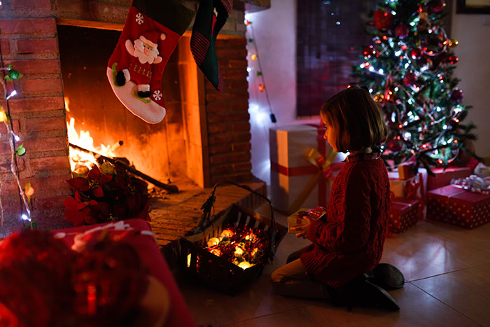 Young child kneeling near a Christmas fireplace with stockings hanging, surrounded by holiday decorations and presents at night.