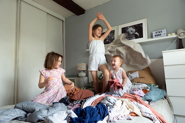 Three children playing on a bed covered with clothes, illustrating a mother lets family live filth to teach a lesson.