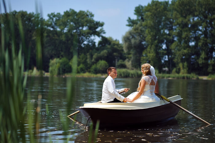 A bride and groom in wedding attire sitting in a small boat on a lake surrounded by trees on a sunny day. - 7