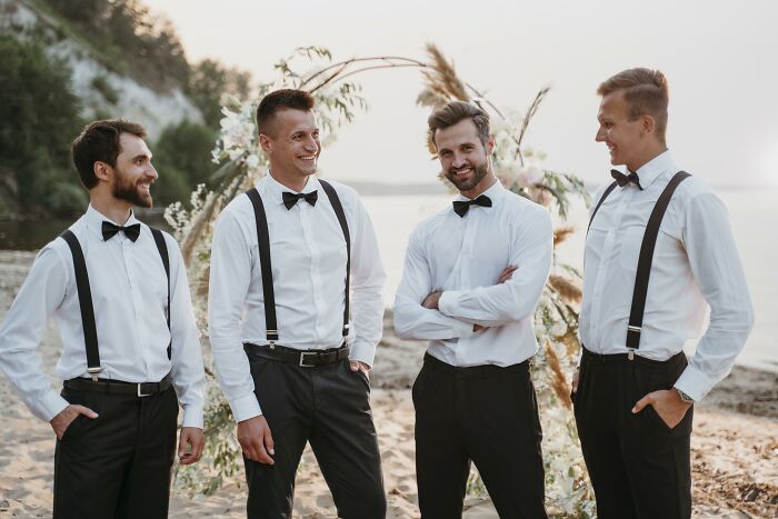 Four groomsmen wearing suspenders and bow ties at an outdoor wedding near a beach, sharing a light moment. - 8