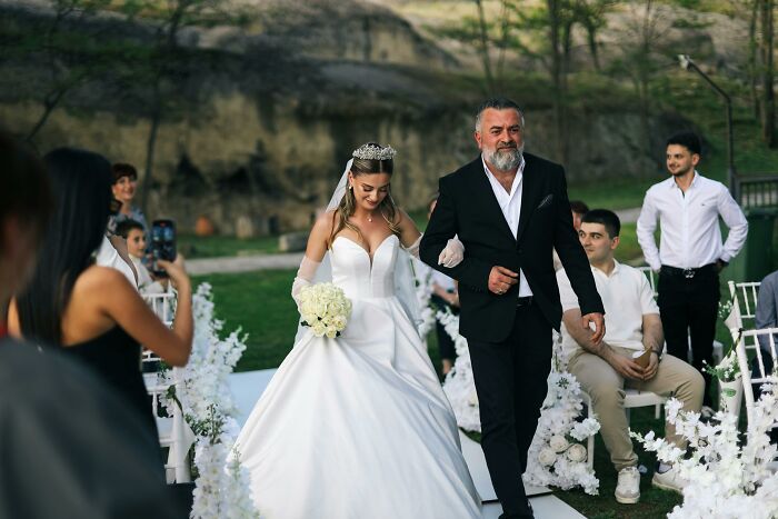 Bride in a white wedding gown walking down the aisle with a man at an outdoor wedding ceremony attended by guests. - 3