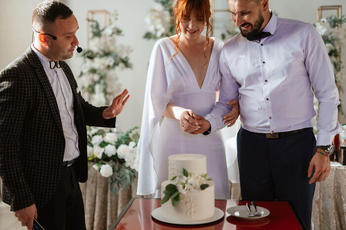Bride and groom cutting wedding cake together, with officiant speaking beside them during wedding celebration. - 22