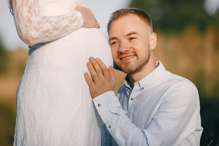 Man smiling and kneeling, gently touching a pregnant woman’s belly at an outdoor wedding celebration. - 13