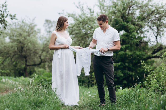 Couple in a green field holding a baby outfit, illustrating stories of attending the same person’s multiple weddings. - 20
