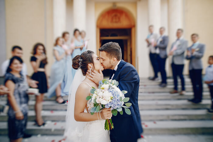 Bride and groom kissing outside a wedding venue with guests watching, illustrating stories of attending multiple weddings. - 11