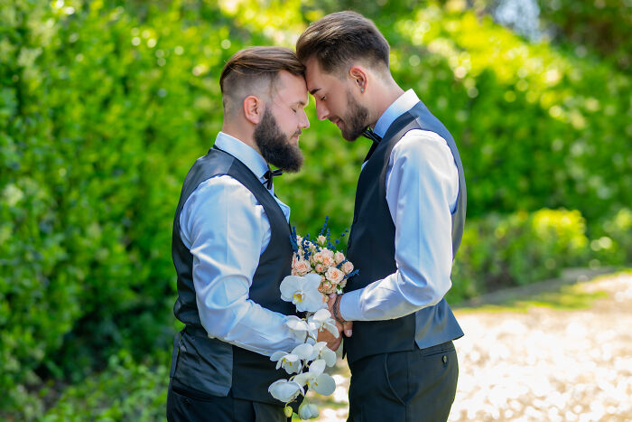 Two grooms in matching vests and shirts share a tender moment at a wedding ceremony outdoors. - 18