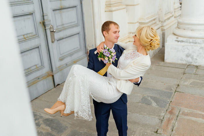 A groom in a navy suit carrying his bride in a white wedding dress holding a bouquet outside a historic building. - 4