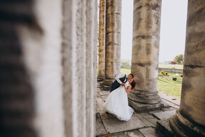 Bride and groom sharing a romantic kiss between large stone columns at a wedding venue during their special day. - 9