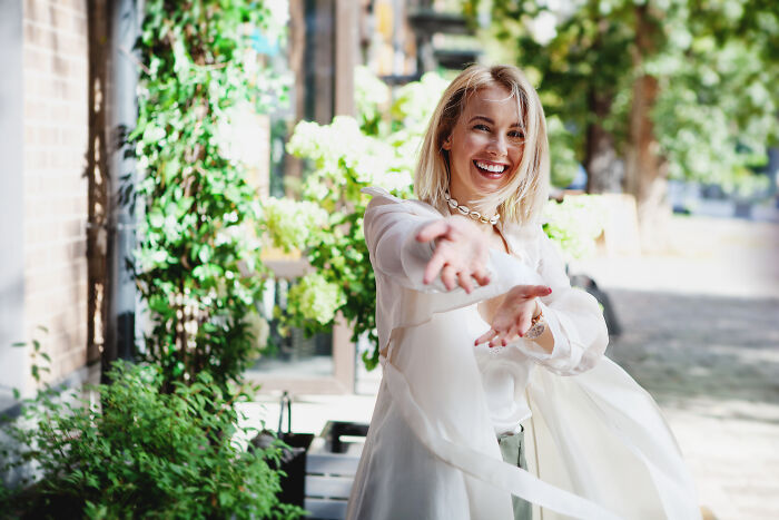 Smiling woman outdoors gesturing with open arms, representing stories about attending the same person’s multiple weddings. - 10