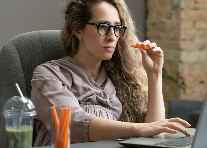 Woman with glasses eating carrot sticks while working on laptop, illustrating marketing campaigns with powerful impact.