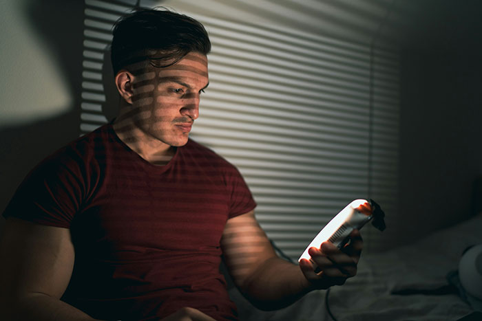 Man looking at phone with shadows of blinds on his face, representing man publicly demoted to friend by GF.