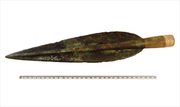 Ancient Neolithic spearhead artifact displayed with measuring scale, linked to groundbreaking discovery under Scottish school halls.