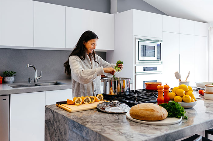 Woman adding fresh herbs to pot in modern kitchen with cooking secrets and ingredients visible on counter. - 2