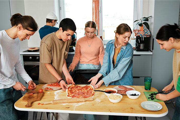 A group of young people preparing pizza together, sharing cooking secrets and unique ingredients in a bright kitchen. - 23