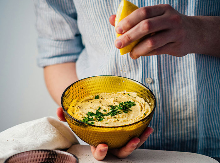 Person squeezing lemon over a bowl of seasoned hummus, sharing cooking secrets and unique ingredients. - 16