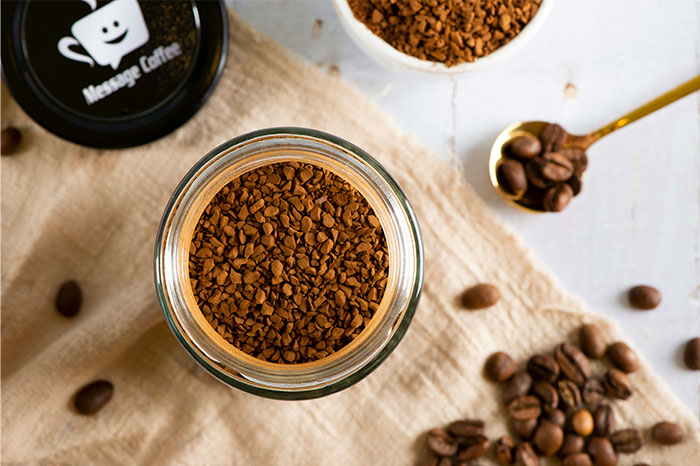 Close-up of instant coffee granules in a jar with scattered coffee beans and a golden spoon on a light fabric surface. - 50