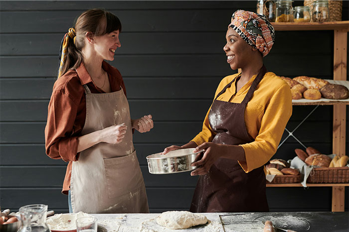 Two women in aprons sharing cooking secrets and ingredients while preparing dough in a rustic kitchen setting. - 6