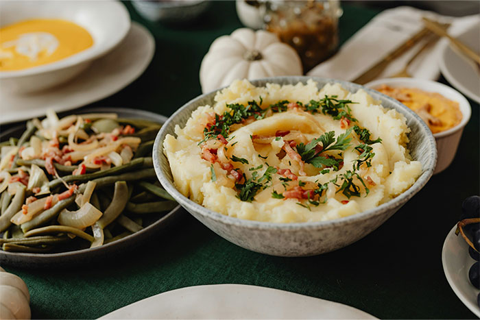 Bowl of mashed potatoes garnished with herbs and bacon, with plates of green beans and soup nearby, sharing cooking secrets. - 25